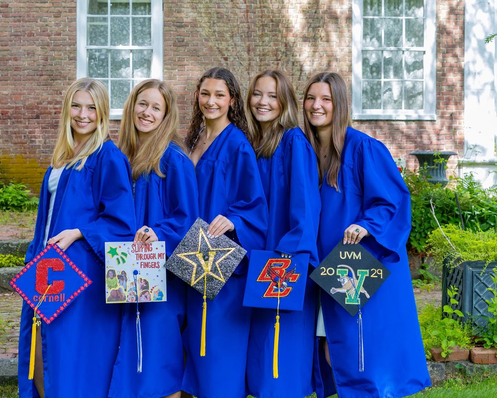 Group of graduates in blue caps and gowns showing decorated graduation caps outside a brick building.