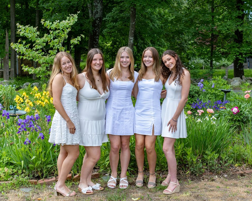Five friends in white summer dresses pose together in a garden with blooming purple iris flowers.