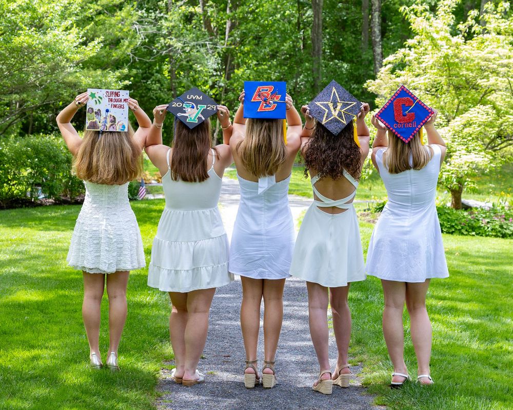 Five graduates in white dresses show decorated caps from behind in garden setting.