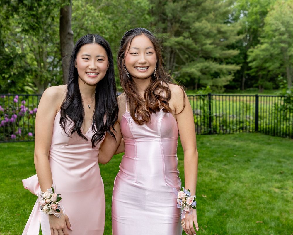 Two friends in matching pink formal dresses pose together on a lawn with a garden fence and evergreen trees in background.