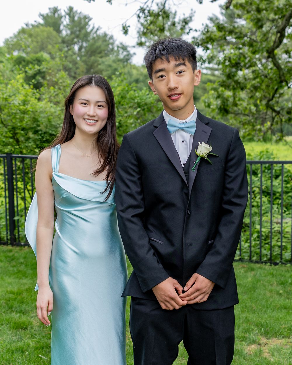 A formal couple stands by a green metal railing outdoors, with her in a satin blue dress and him in a black tuxedo.