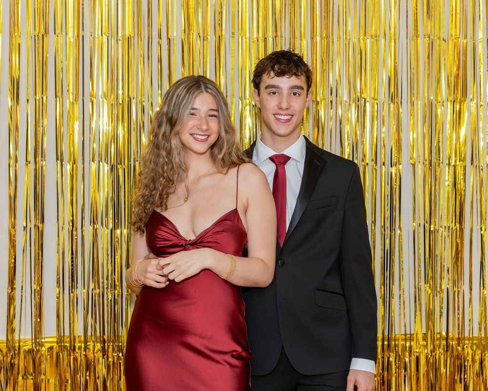 A couple poses together at a formal event against a shimmering gold metallic streamer backdrop.
