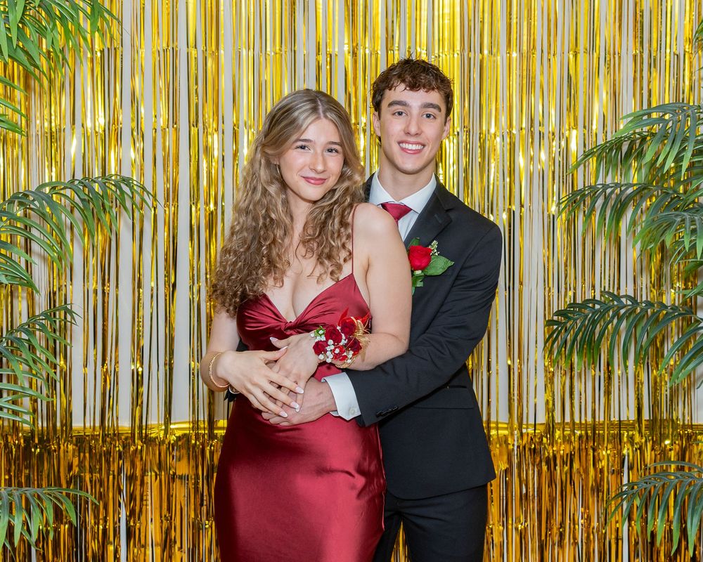A couple poses at a school dance or prom in front of a gold foil backdrop with palm plants.