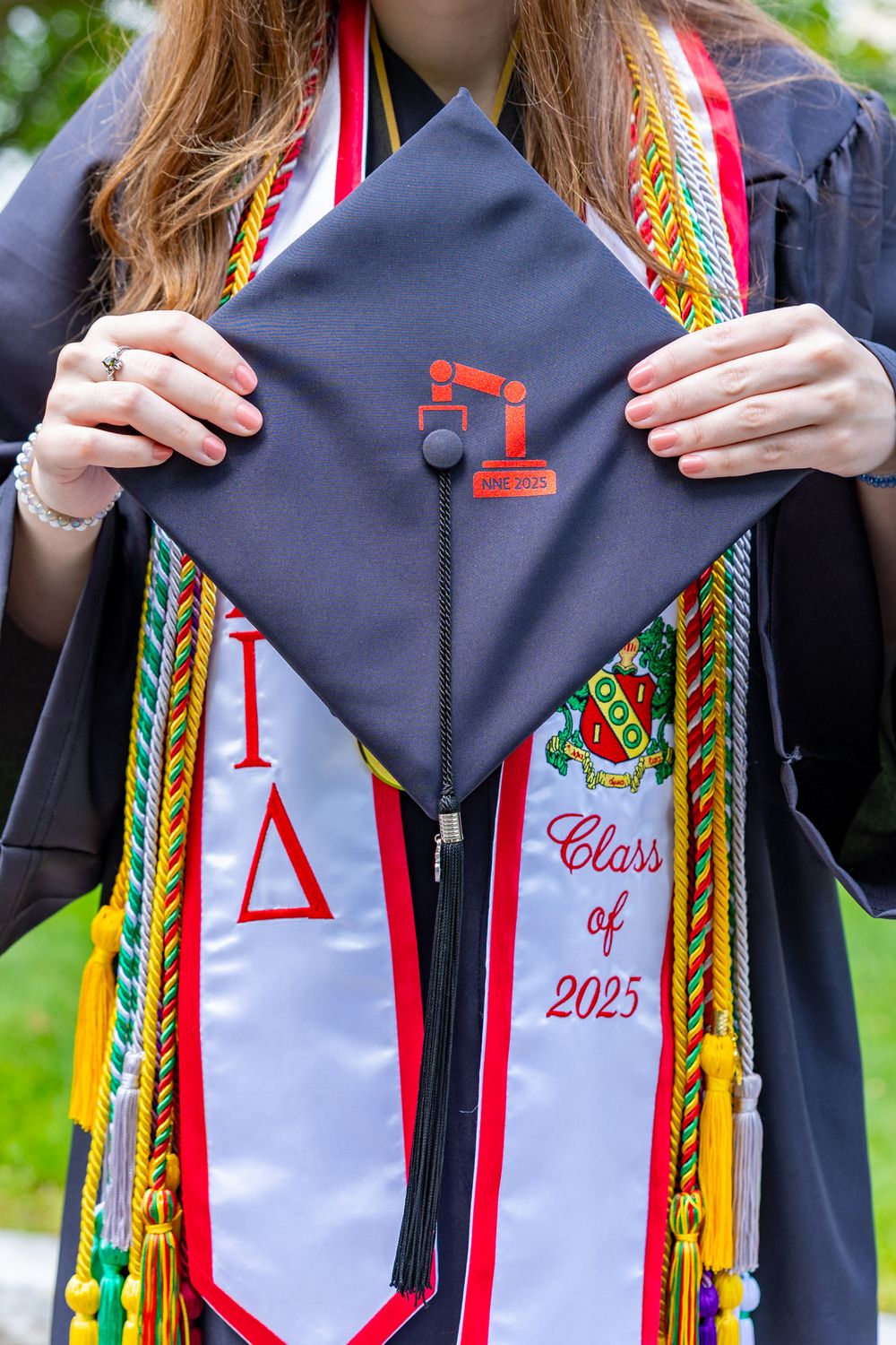 Graduate in academic regalia wearing colorful cords and stoles poses outdoors near blooming white flowers. A cap and gown graduate portrait.
