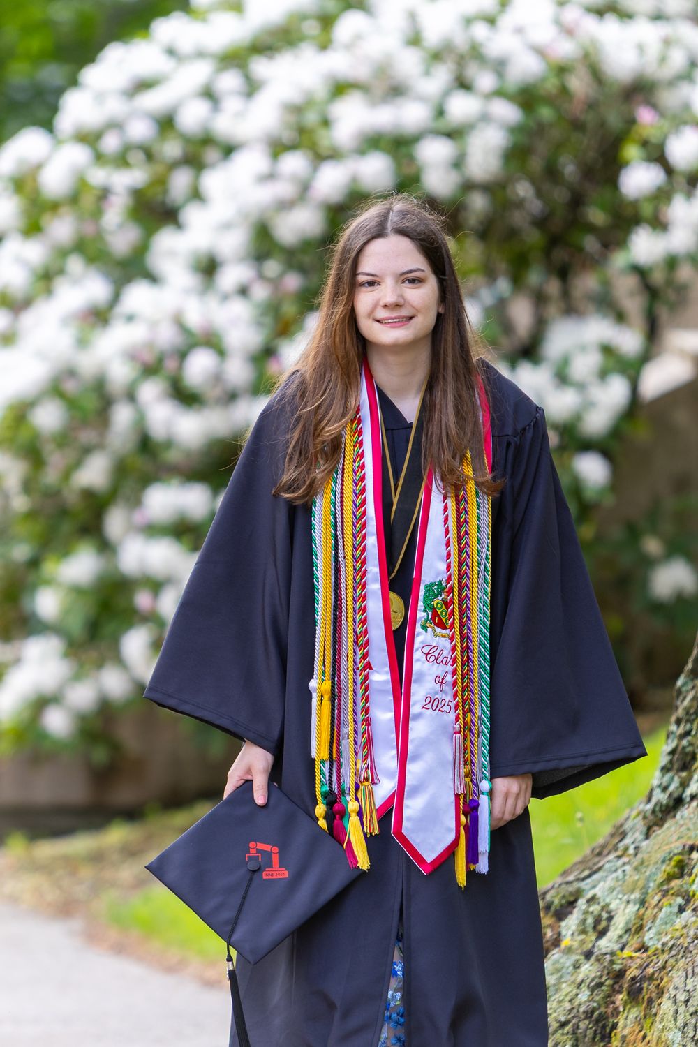 Graduate in academic regalia wearing colorful cords and stoles poses outdoors near blooming white flowers. A cap and gown graduate portrait.