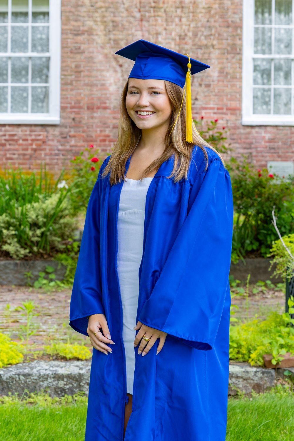 Graduate in blue cap and gown poses outdoors in front of brick building.