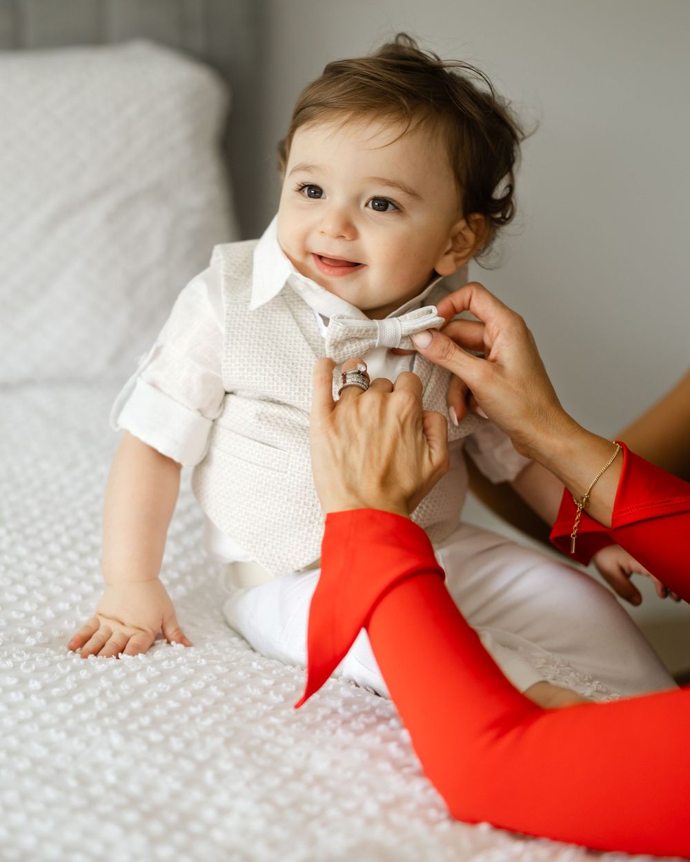 A baby in a white formal outfit with a bowtie sitting on a bed.
