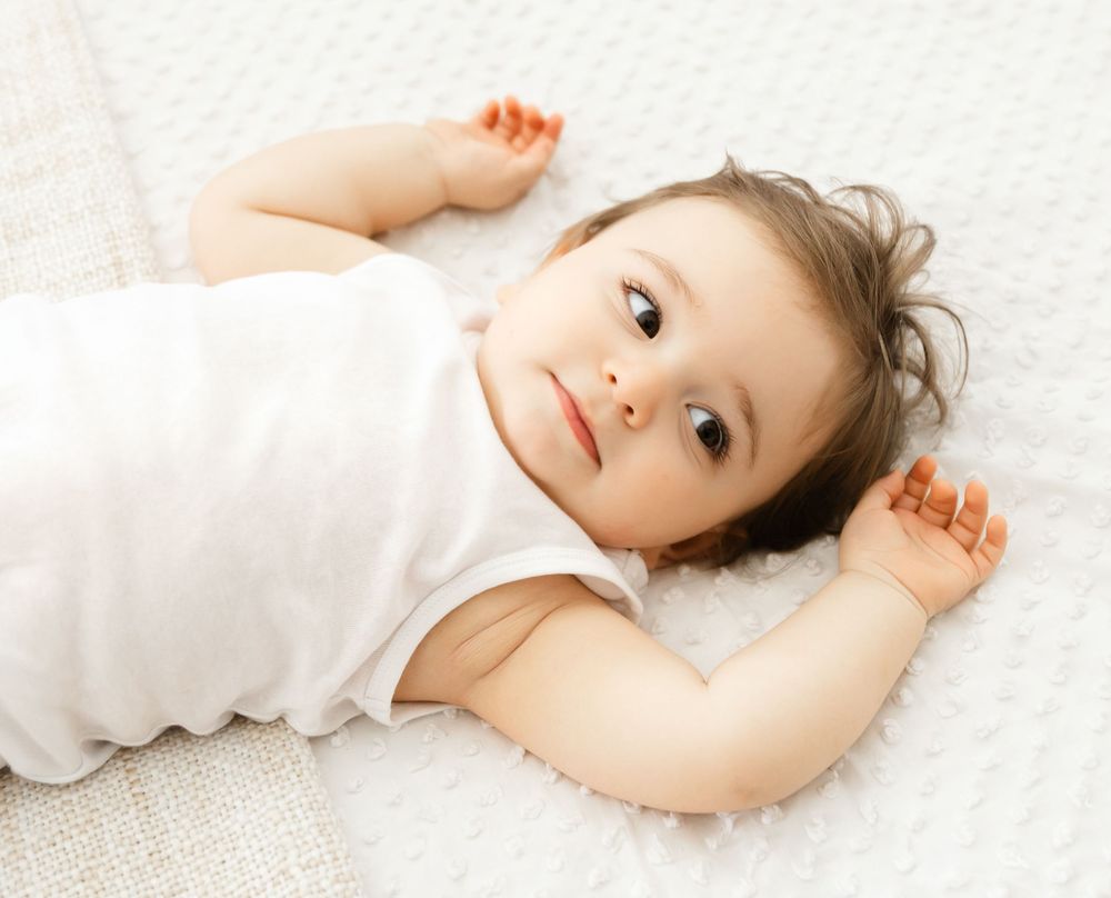 Baby in white onesie lying on back on soft white carpet.