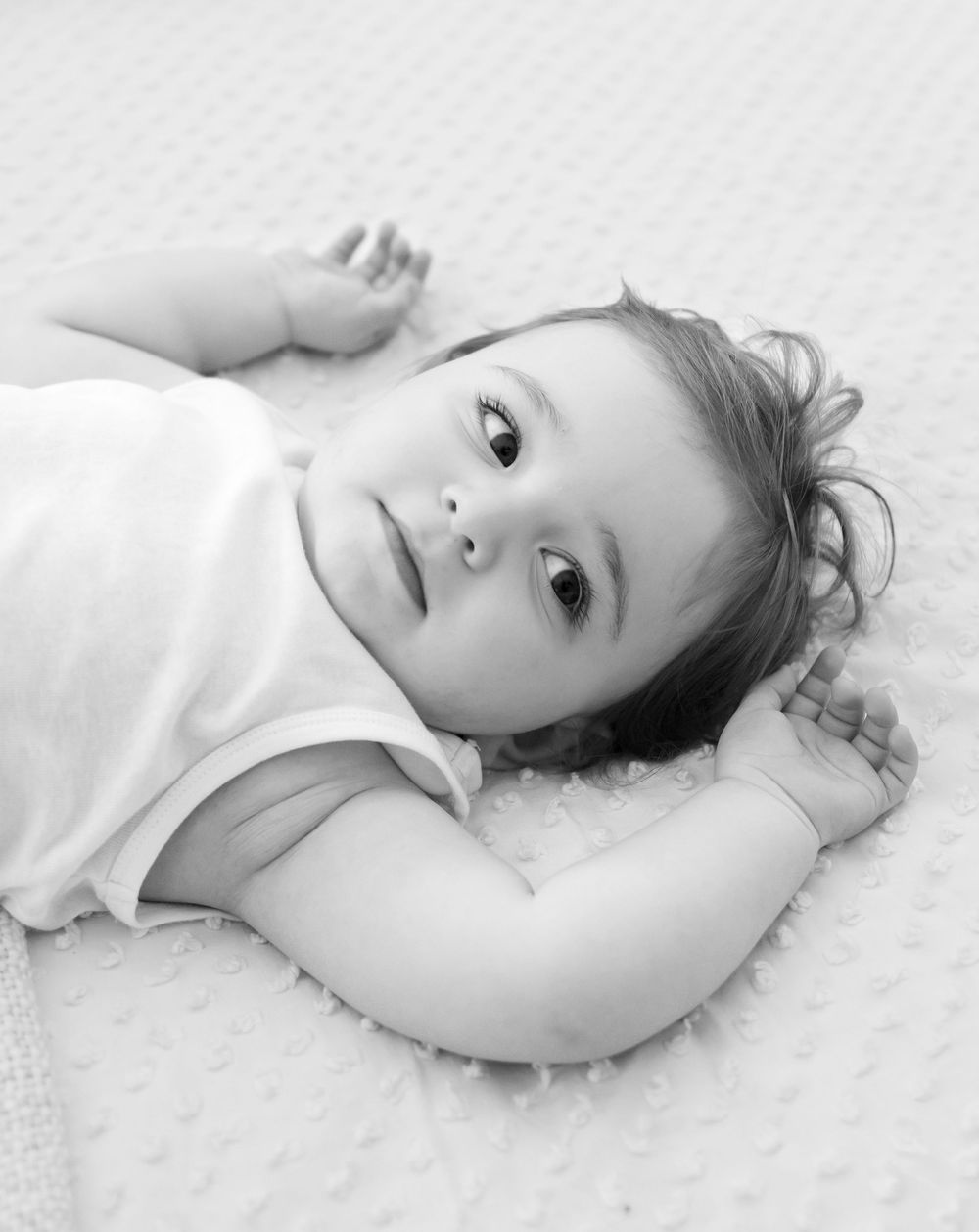 A black and white portrait of a young toddler lying on white fabric looking up at the camera.