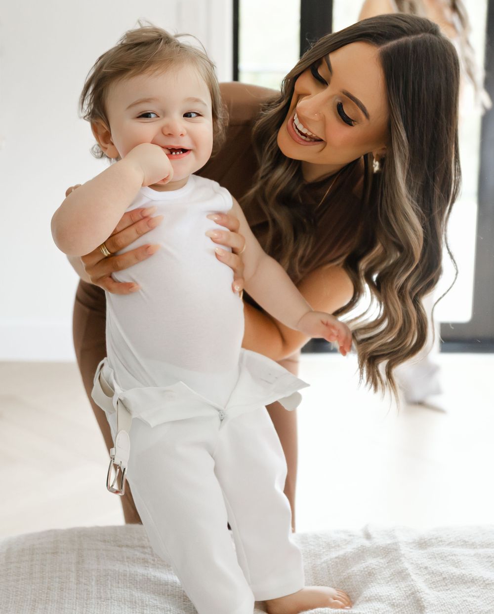 Mother and baby in white outfits share tender moments together while sitting on a bed.