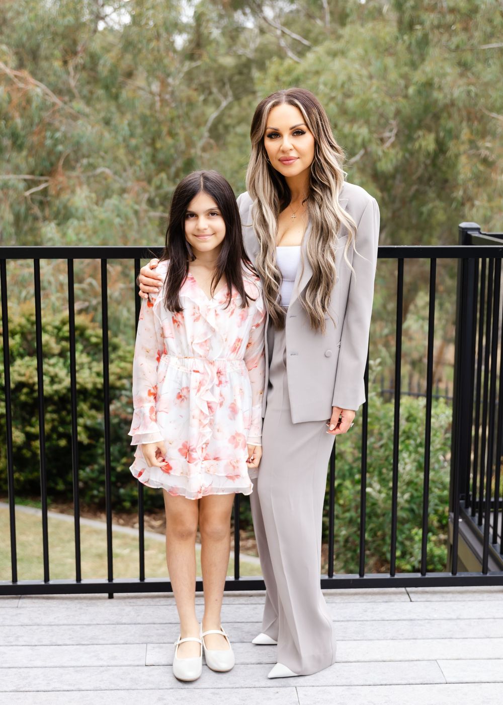 Two people in elegant outfits standing together on outdoor deck with greenery in background.