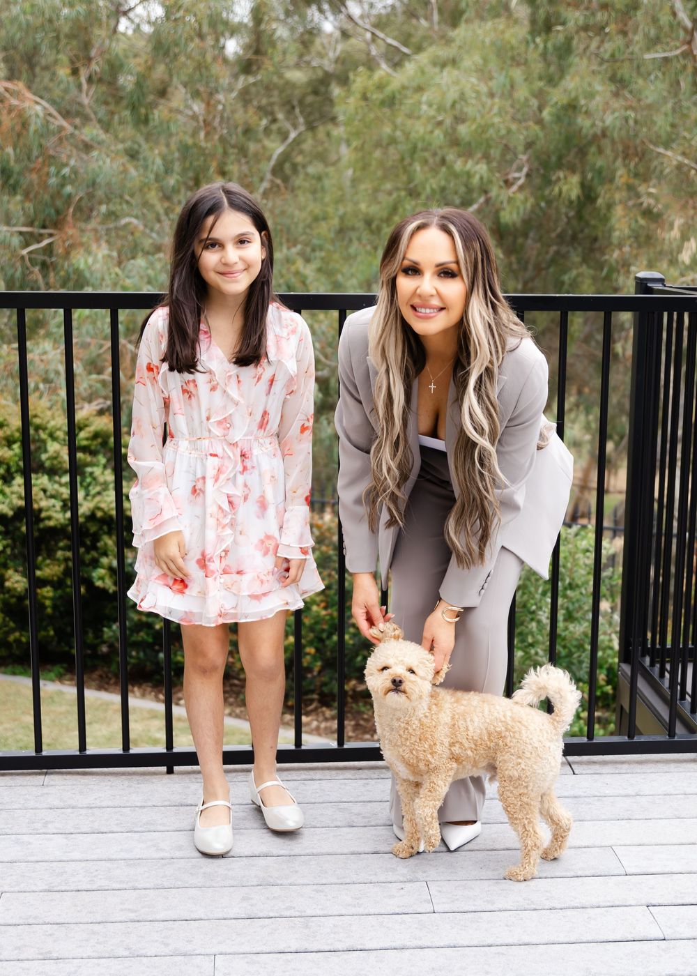 Two companions posing together with a small fluffy tan dog on a wooden deck with a black railing and natural backdrop.
