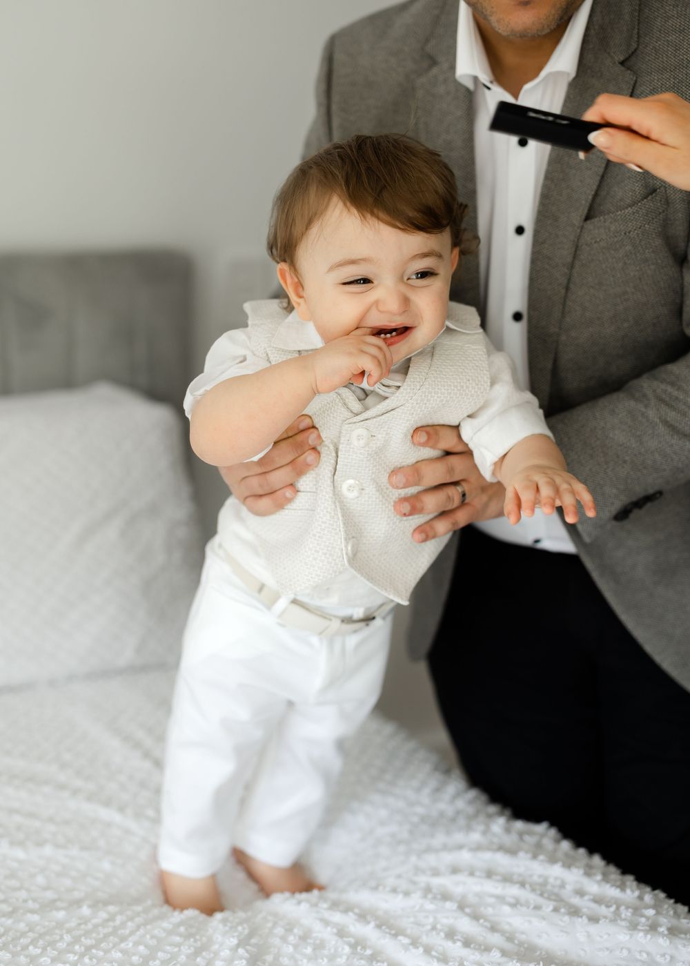 A happy baby wearing all white formal attire standing on a bed.