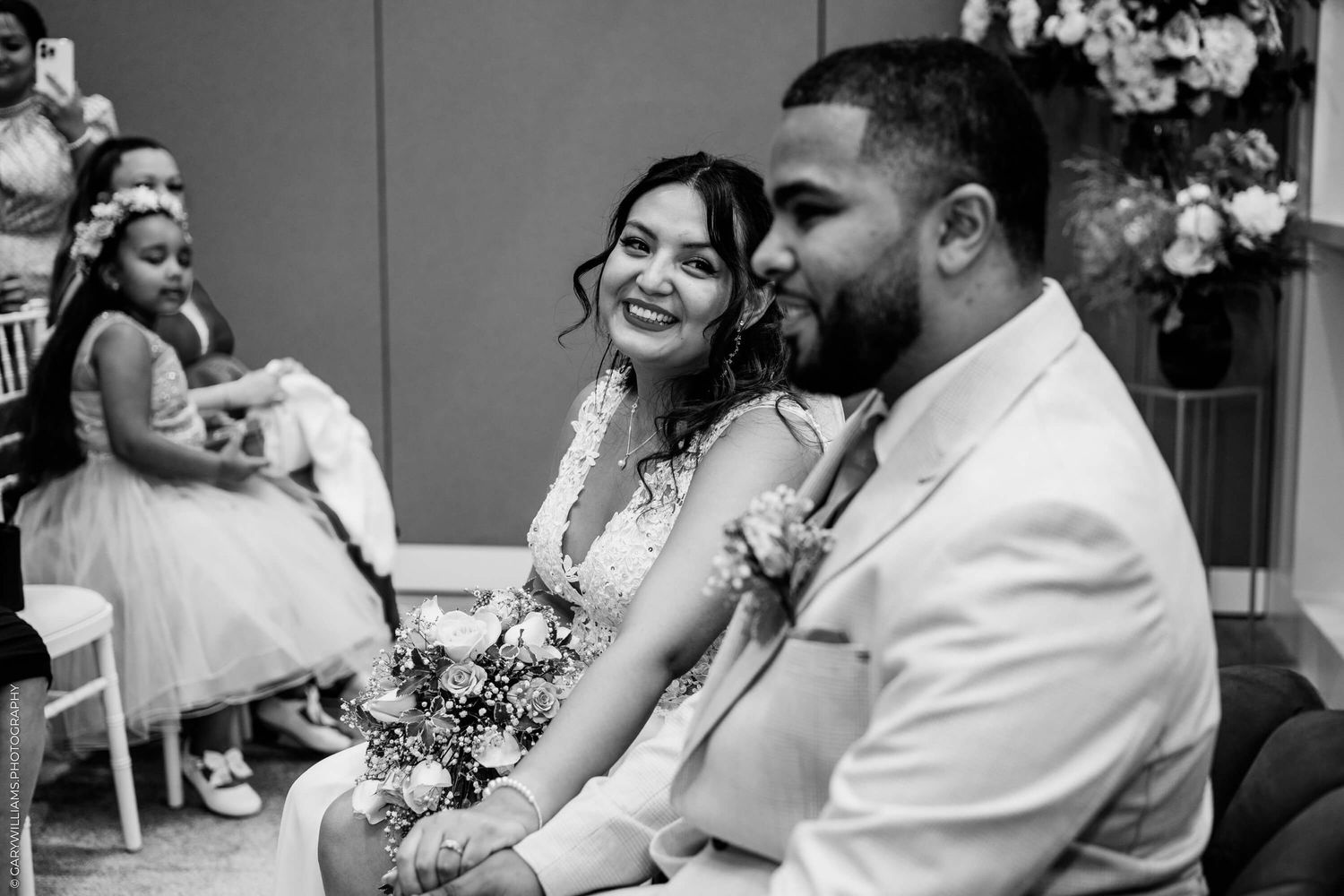 A candid black and white moment at a wedding ceremony shows smiling couple sitting together with floral decor in the background.