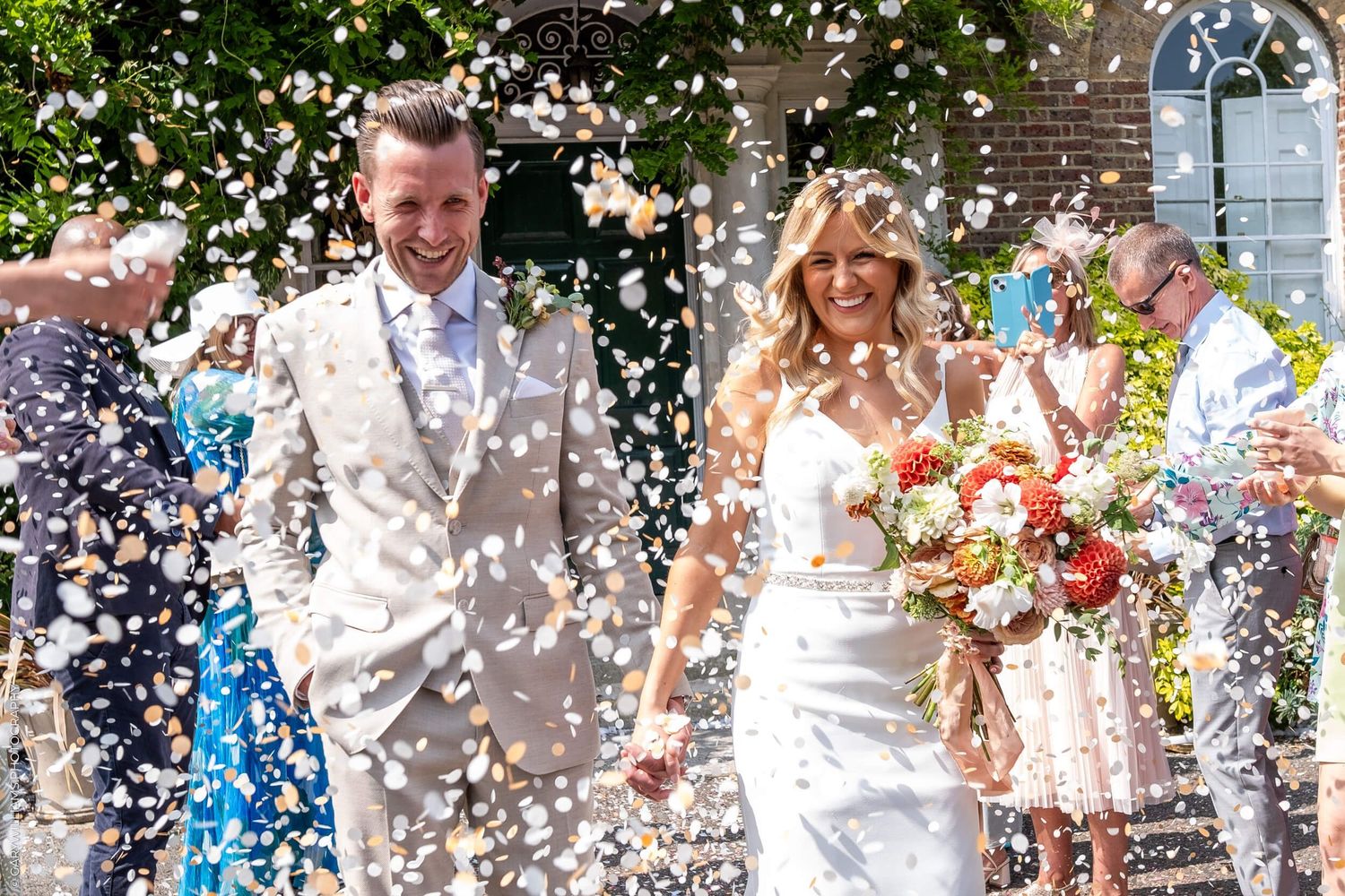 A newlywed couple exit their wedding ceremony under a shower of white confetti on a sunny day outside a church.