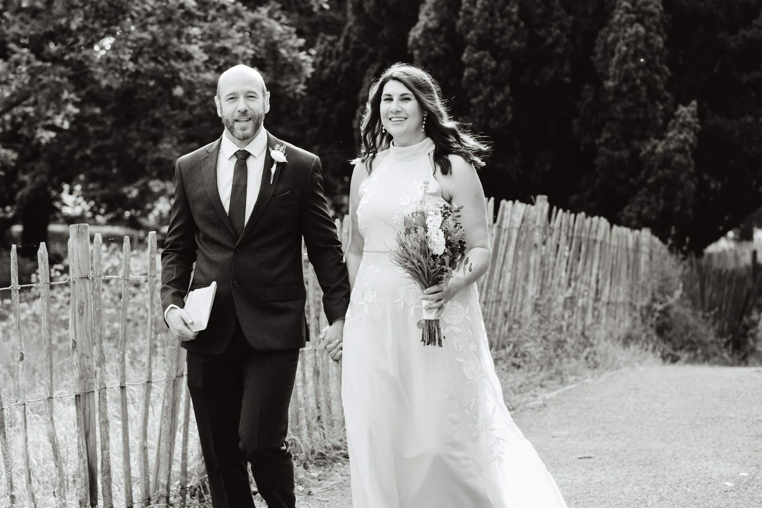 A bride and groom walk together along a rustic path with wooden fencing in this black and white wedding photo.