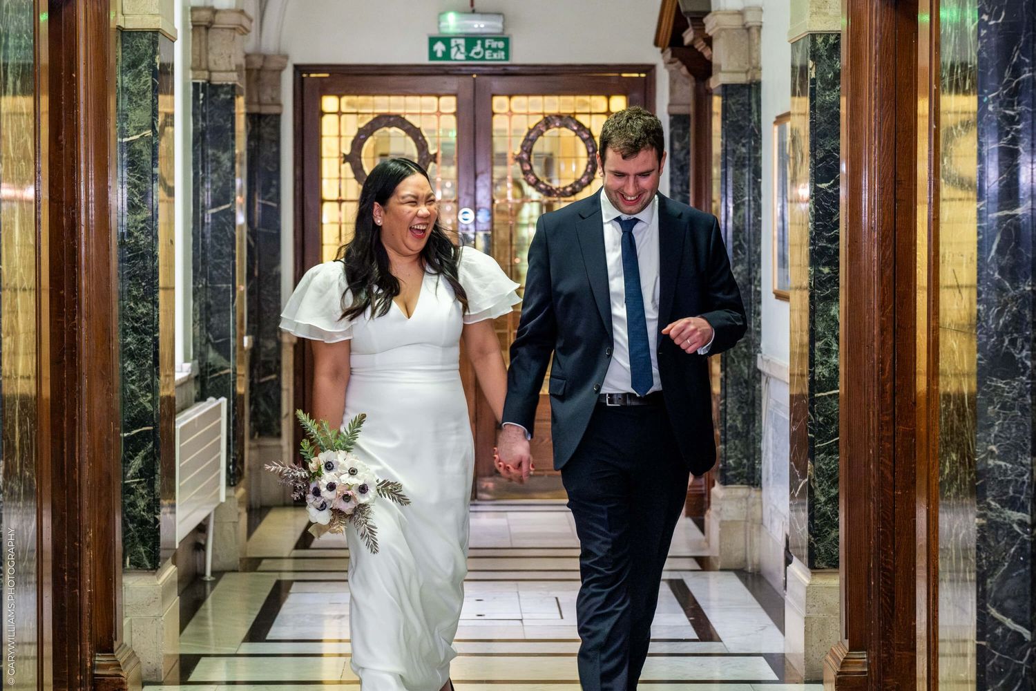 Two people walking down an elegant marble hallway with ornate display cases and an exit sign visible.