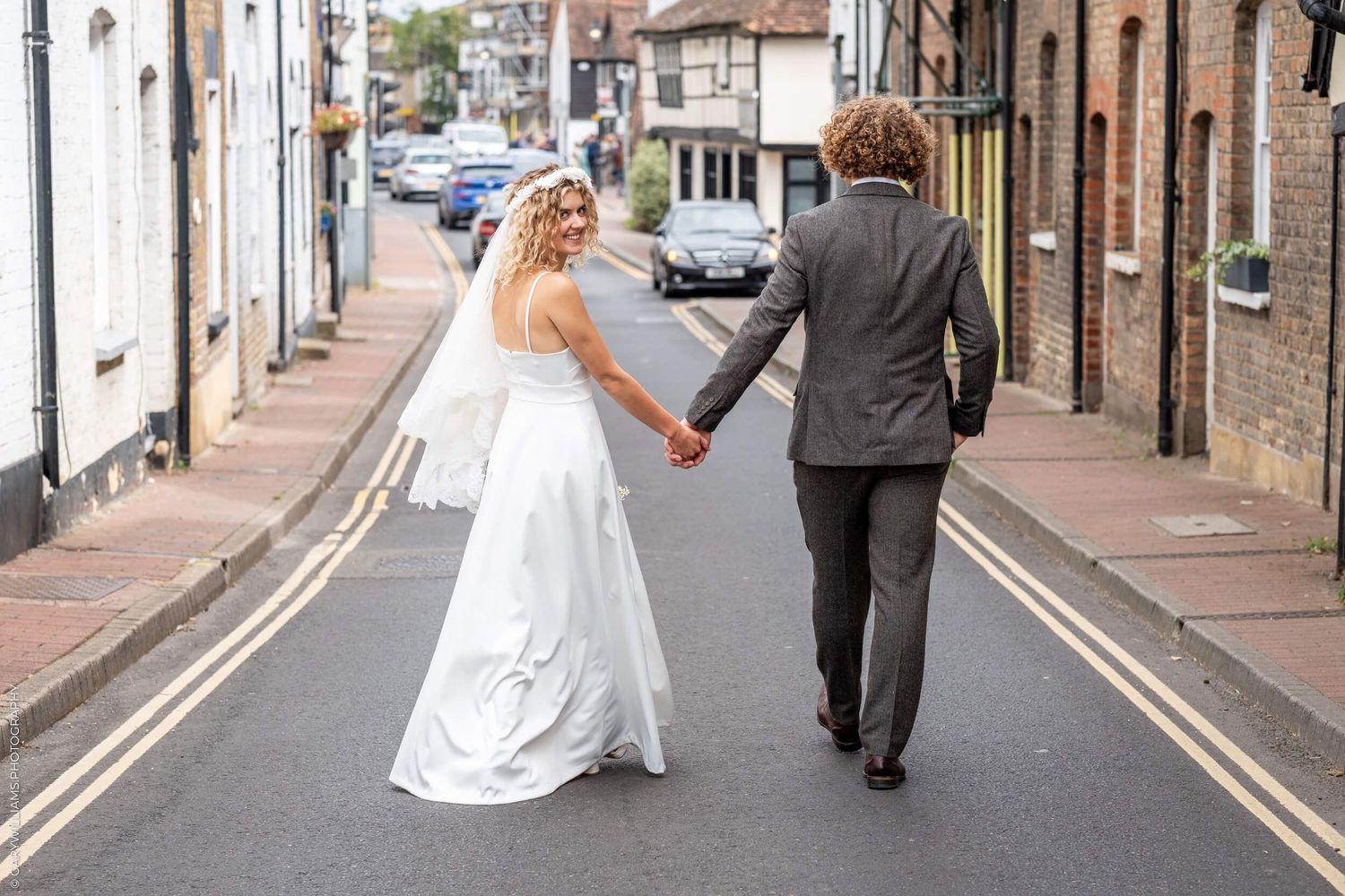 Back view of newlyweds holding hands while walking down a British street with vintage buildings.