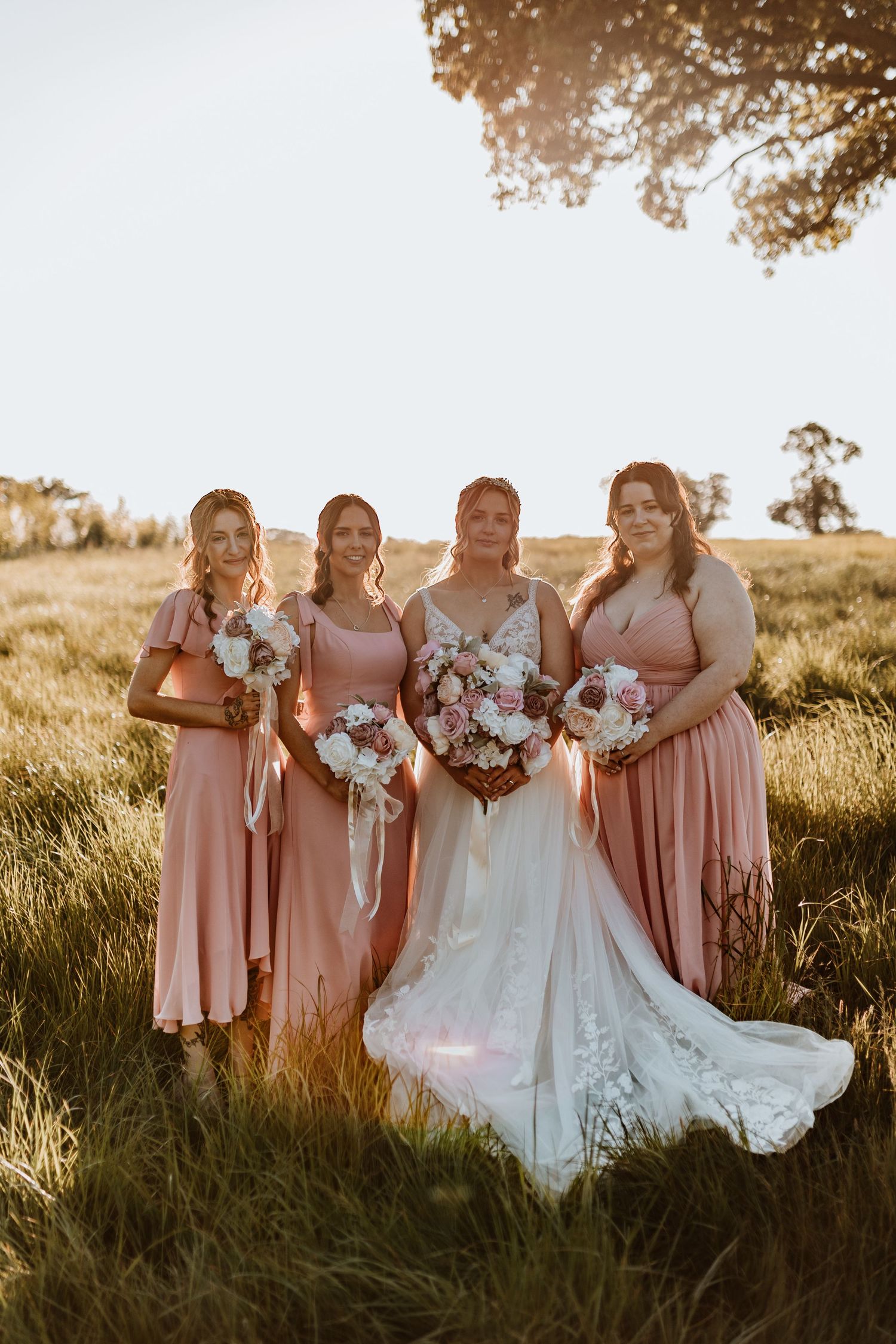 A bridal party in blush pink dresses poses in a sunlit meadow field holding white floral bouquets at Stanford Farm, Shropshire.