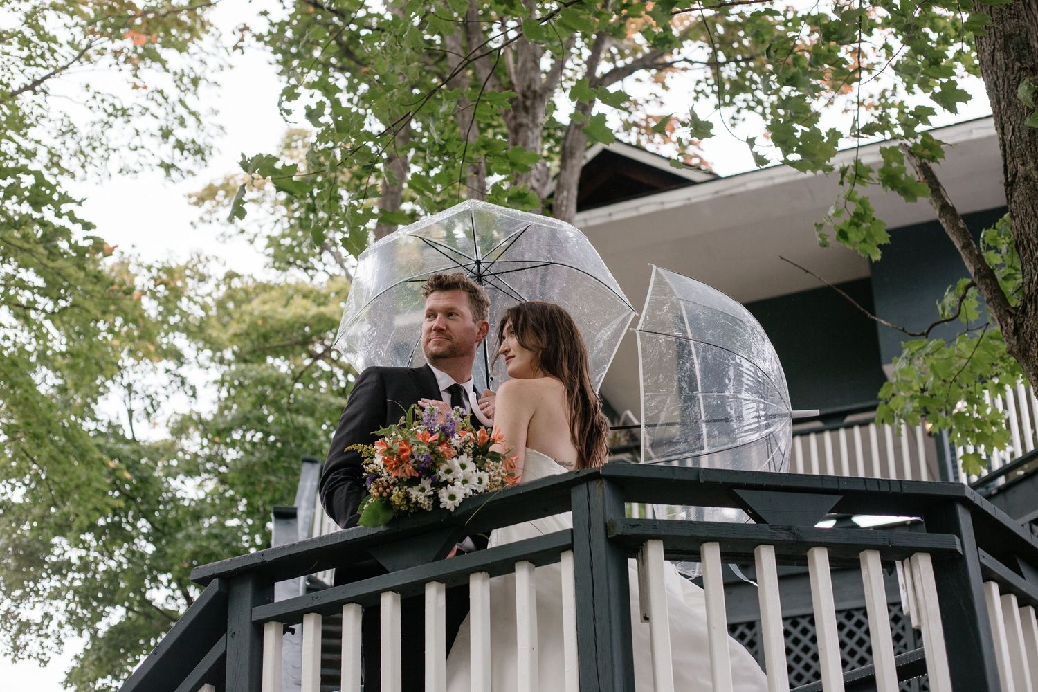 A couple stands together on a wooden balcony under a clear umbrella surrounded by green trees on a rainy day.