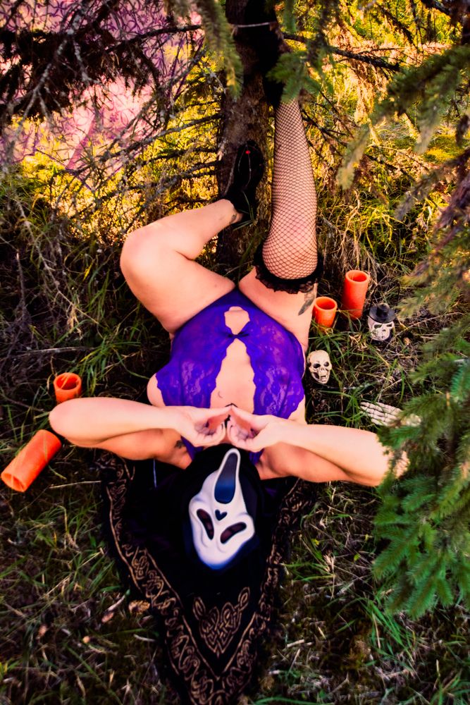 Gothic Halloween scene with purple outfit, fishnet stockings, mask, and spooky decorations on forest floor.