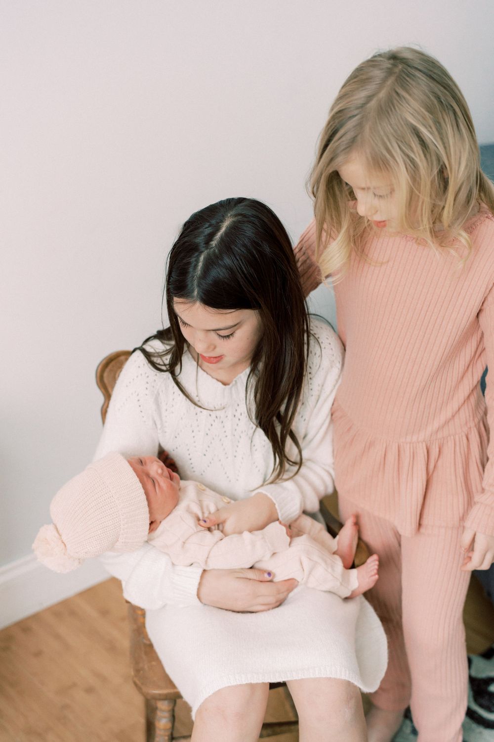 Children gathered around a baby wearing pink clothing while sitting indoors.