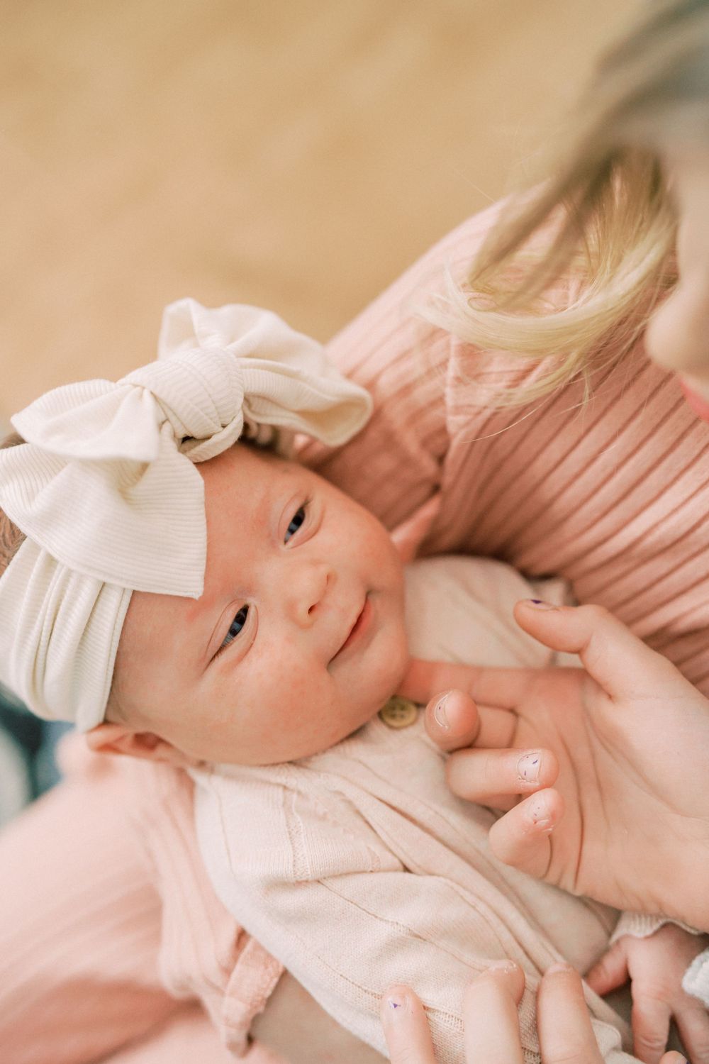 Baby wearing white bow headband smiling while lying in soft pink clothing.