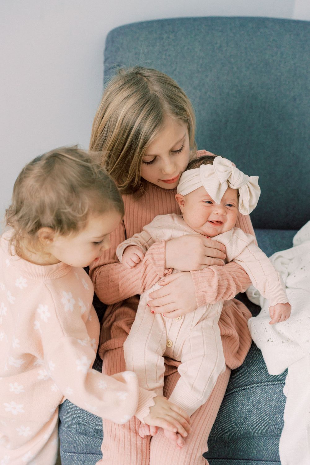 Three siblings sharing tender moment on teal armchair in coordinated pink outfits.