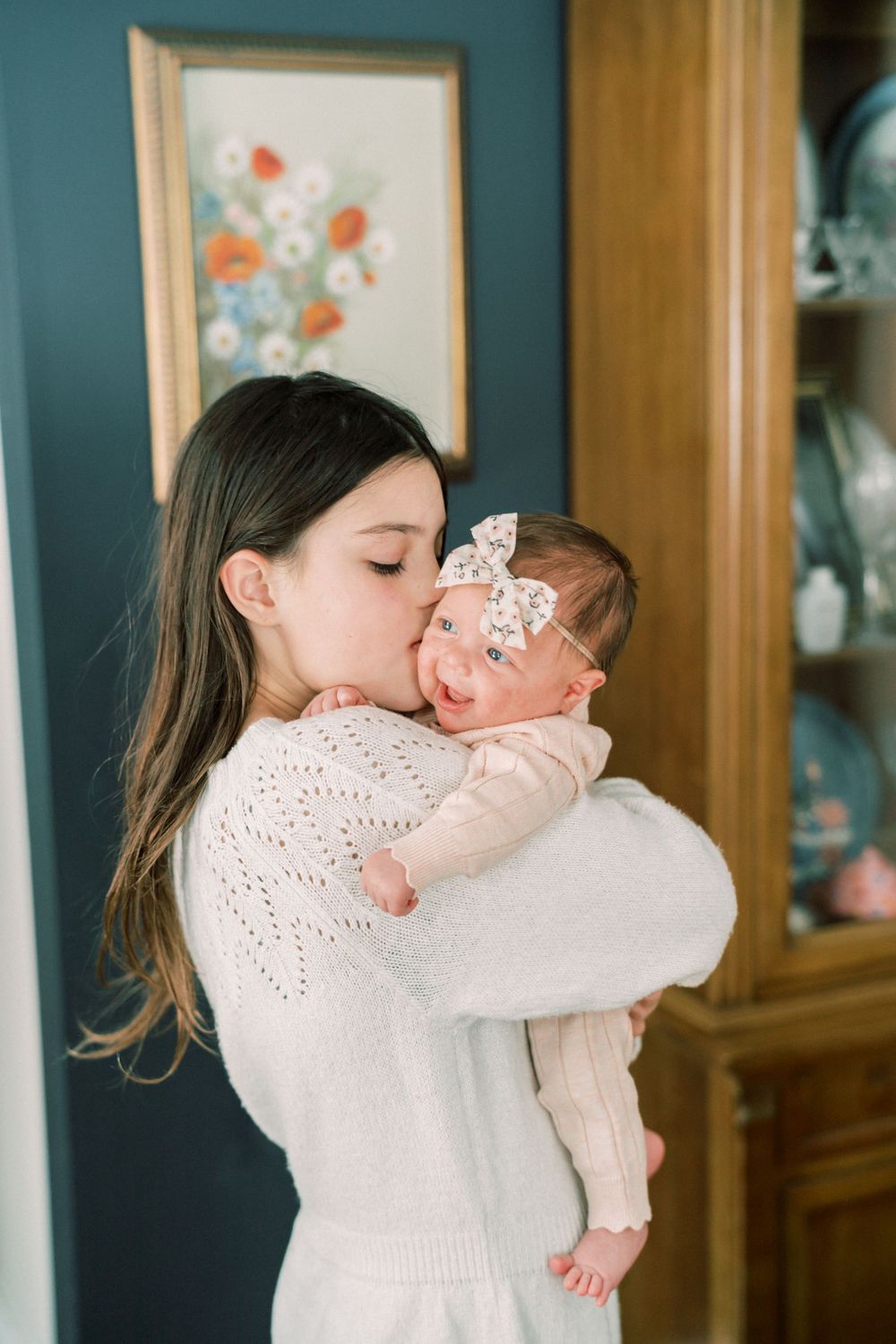 Woman in white sweater kisses baby wearing lace headband against blue wall with floral art.