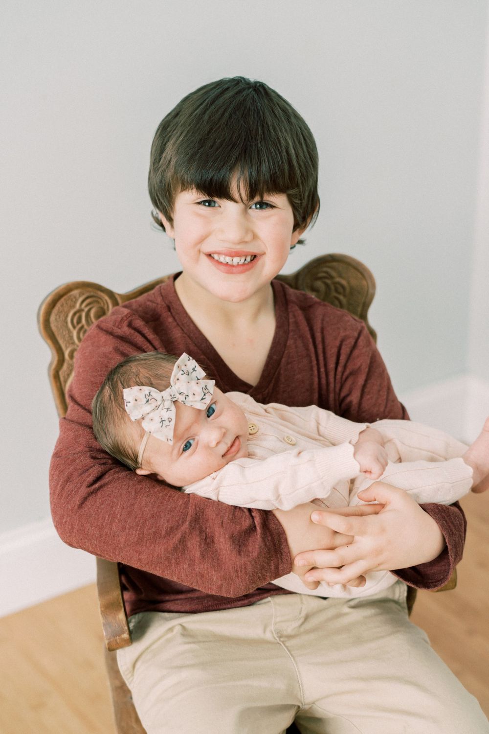 Young boy in brown sweater holds baby wearing bow while sitting in wooden chair.