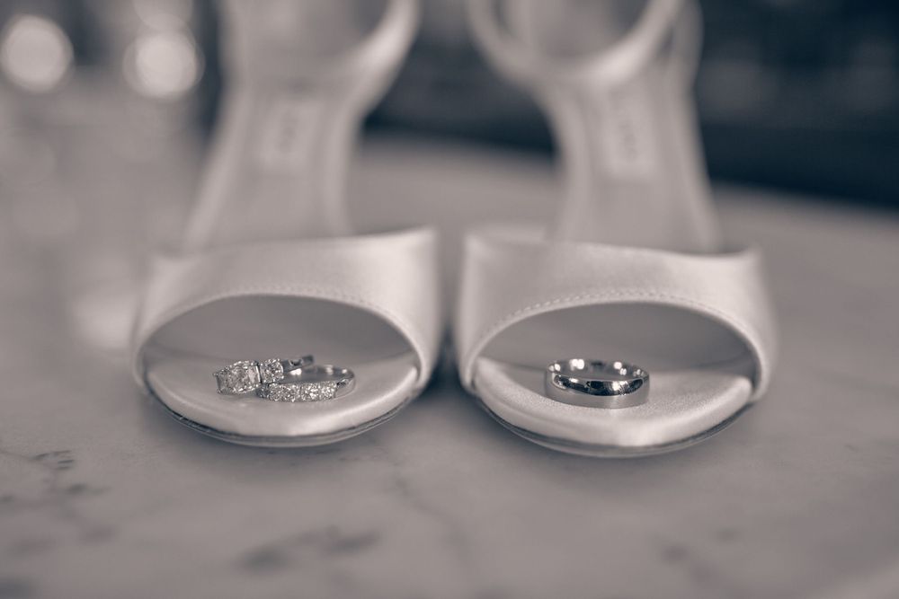 Wedding rings placed on white peep-toe bridal shoes in a soft black and white photograph.