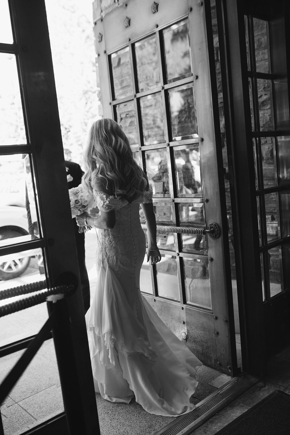 A bride in a flowing white lace gown stands silhouetted against a vintage windowed door in black and white photography.