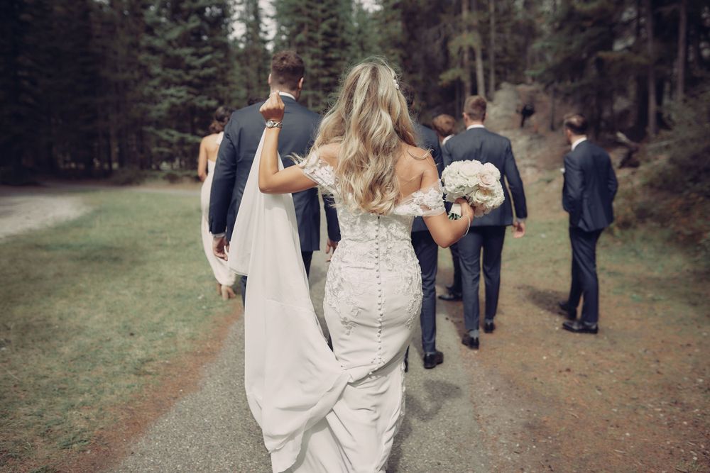 A group walks along a forest path as a wedding party, with the bride's detailed white lace dress visible from behind Banff wedding photo