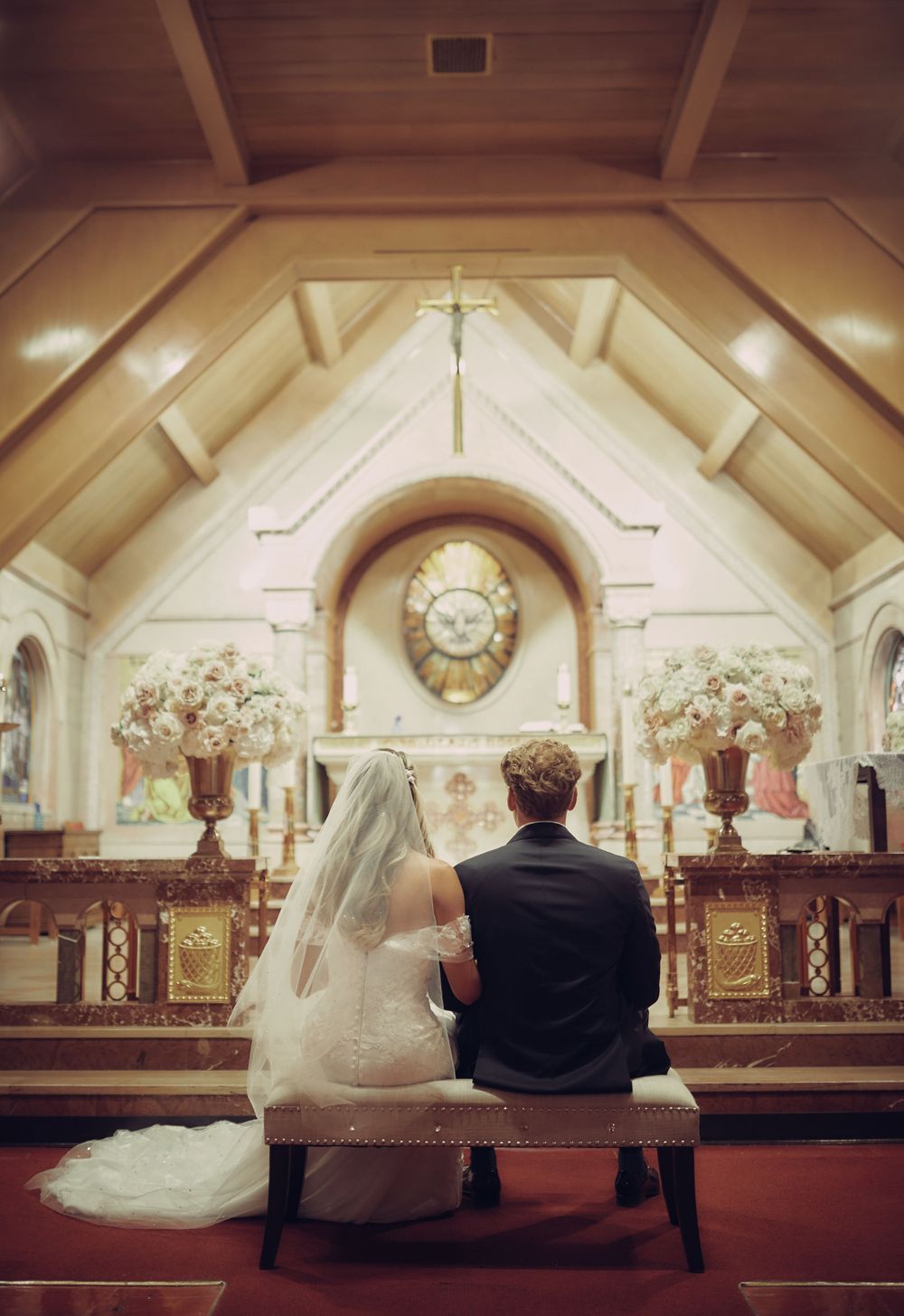 A couple sits on a bench inside an ornate church sanctuary with vaulted ceilings and floral arrangements at the altar of a Catholic church