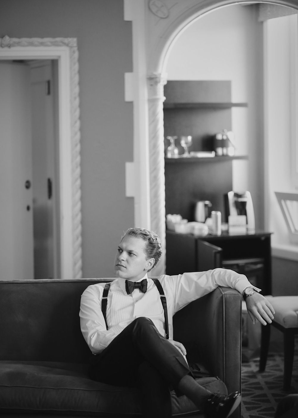 Vintage black and white portrait of a well-dressed groom in bow tie and suspenders lounging on a dark leather couch.