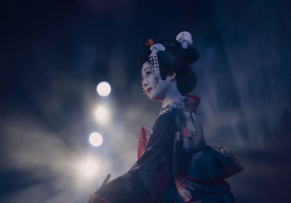 A Japanese woman in an elaborate kimono silhouetted against a dark moody background with soft bokeh lights.