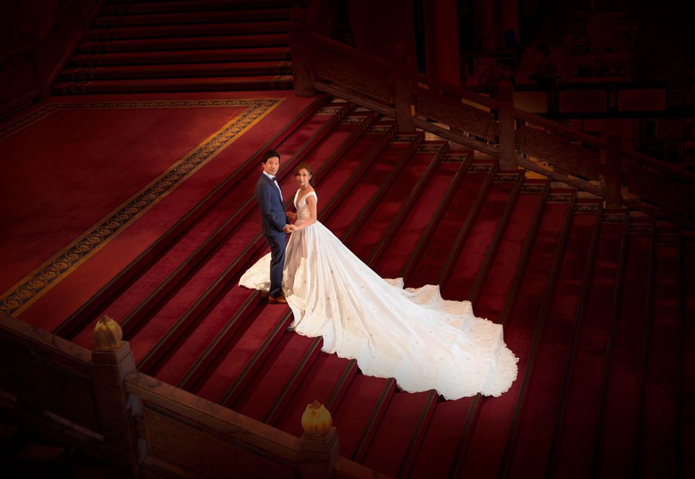A wedding couple stands on elegant red carpeted stairs in a grand theater, with a flowing white gown trailing behind them. International