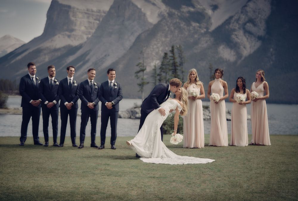 A wedding party in navy suits and beige dresses stands against a backdrop of mountains and lake in Banff, Canada. Banff wedding photographer