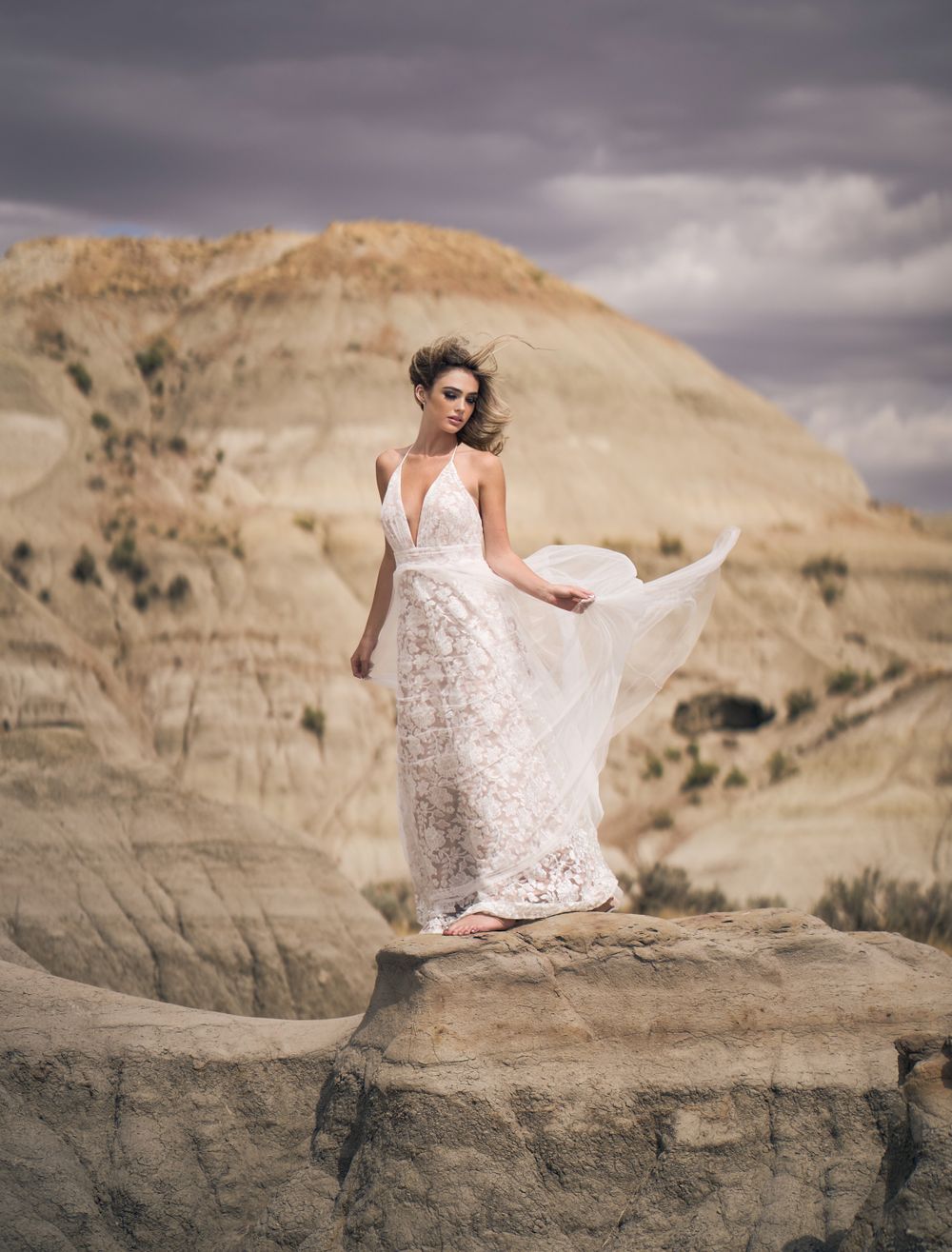 A person in a flowing white lace wedding dress stands elegantly on a rocky ledge against dramatic desert cliffs and stormy skies.