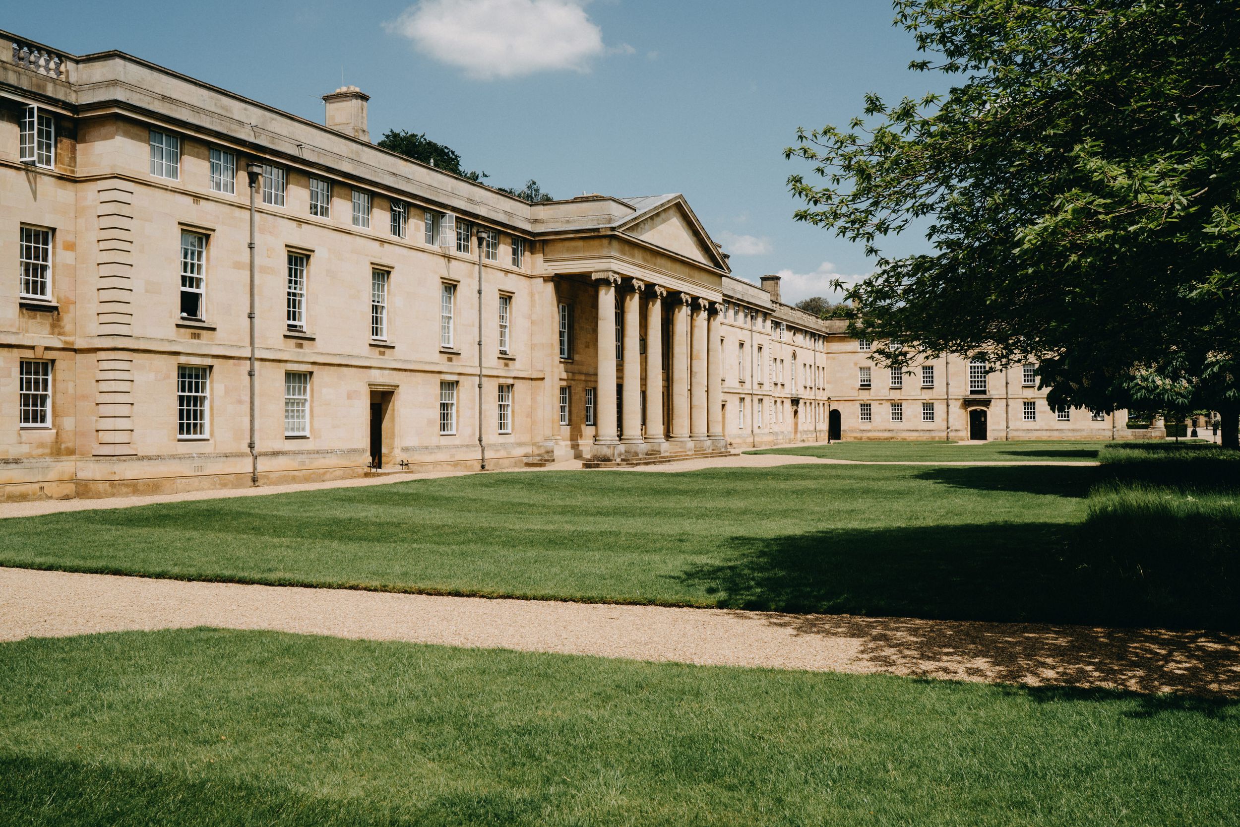 Downing College Cambridge - grand historic stone building with columned entrance and manicured lawn on sunny day.