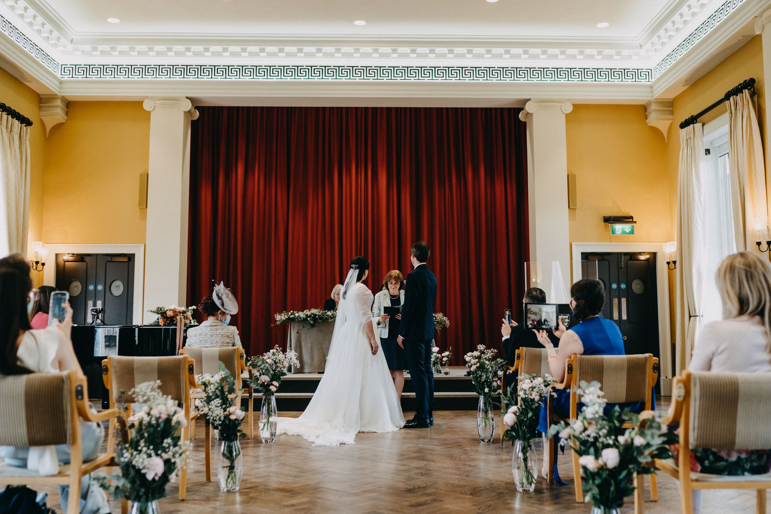 An indoor wedding ceremony at Downing College Cambridge with red curtains and floral aisle decorations in an elegant hall.