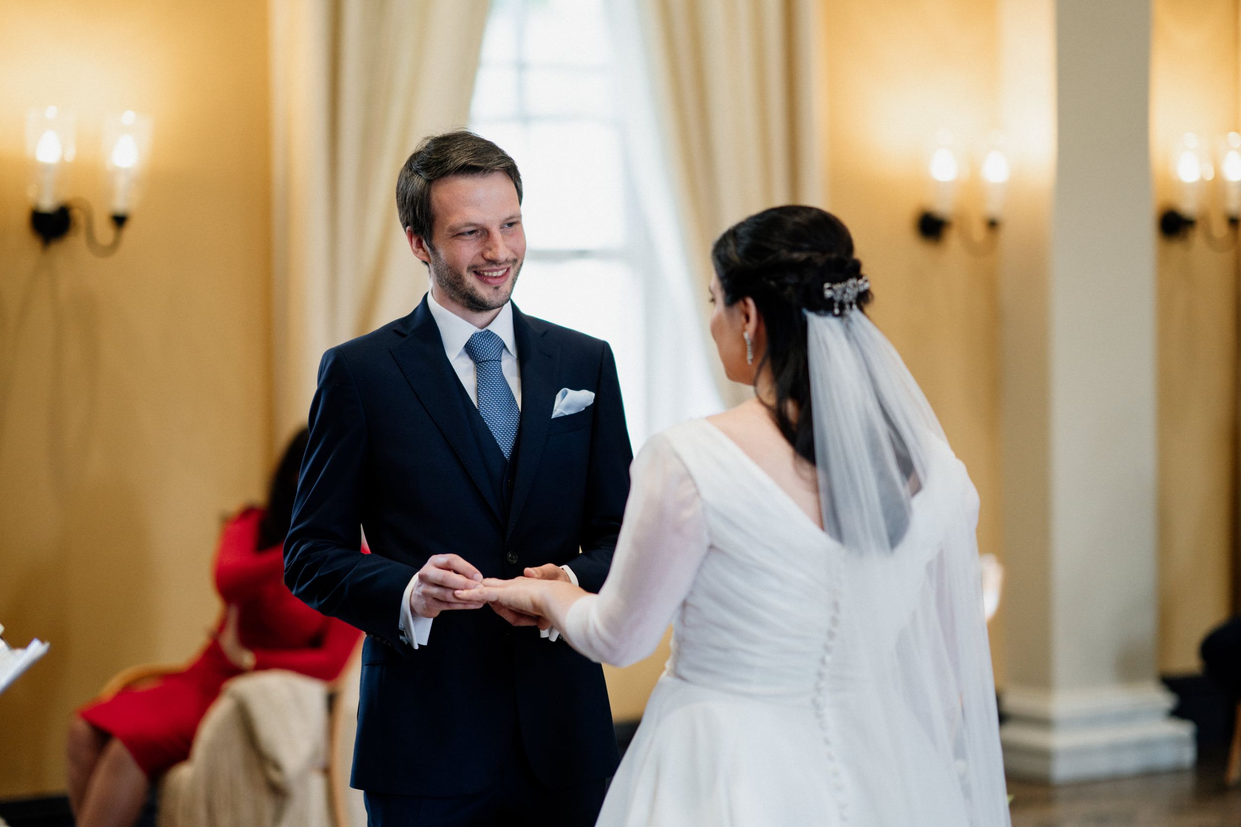 A joyful wedding ceremony exchange of rings between bride and groom in an elegant room with yellow walls and sconces.