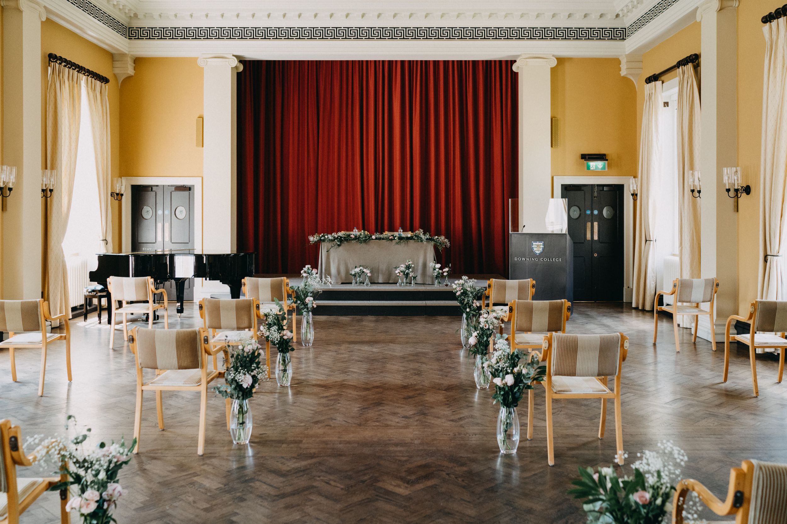 Elegant ceremony room setup with floral arrangements and wooden chairs facing red curtained stage at Downing College Cambridge.