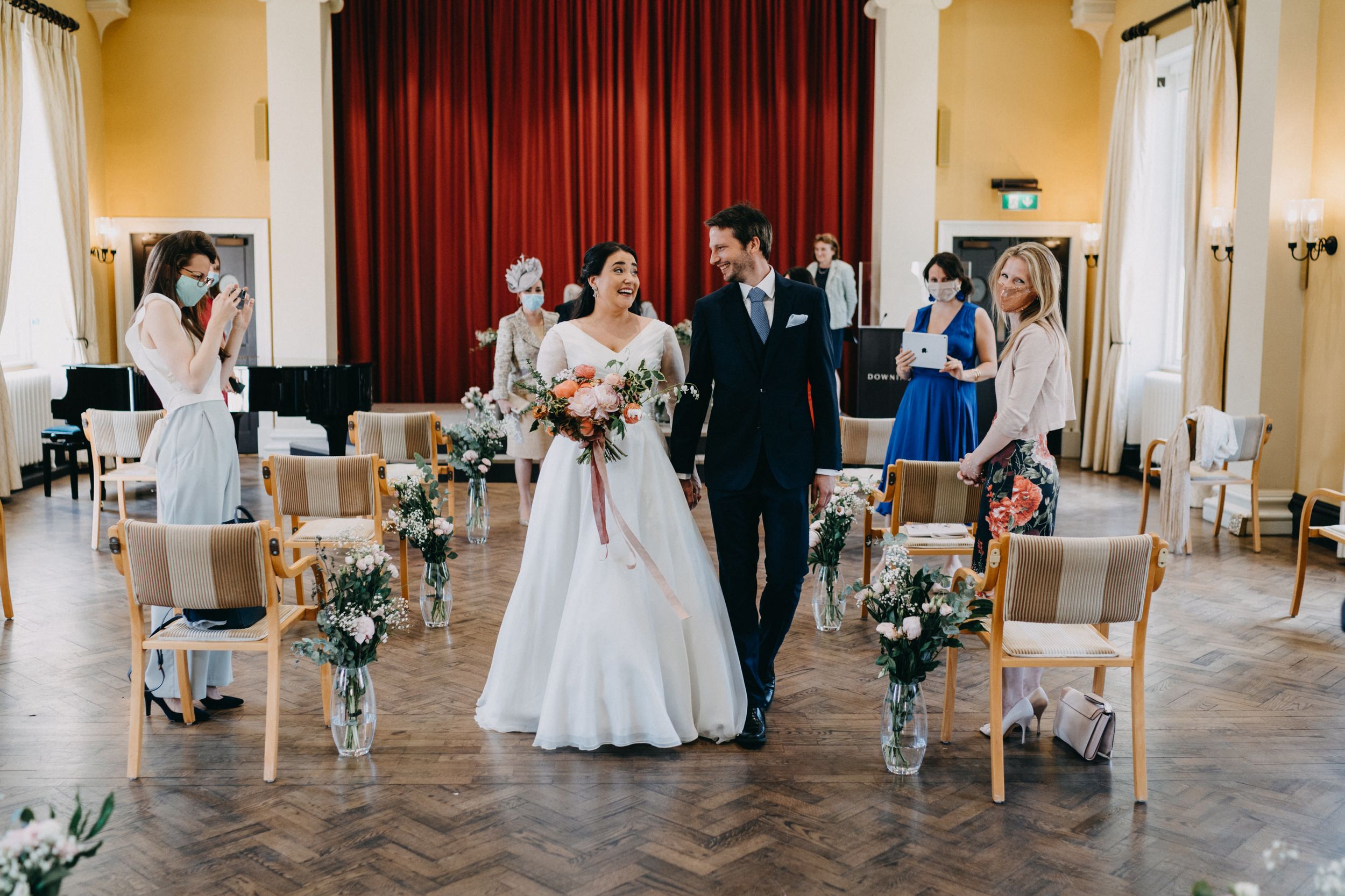 Wedding ceremony at Downing College, Cambridge in an elegant room with red curtains and floral-decorated white chairs lining the aisle.