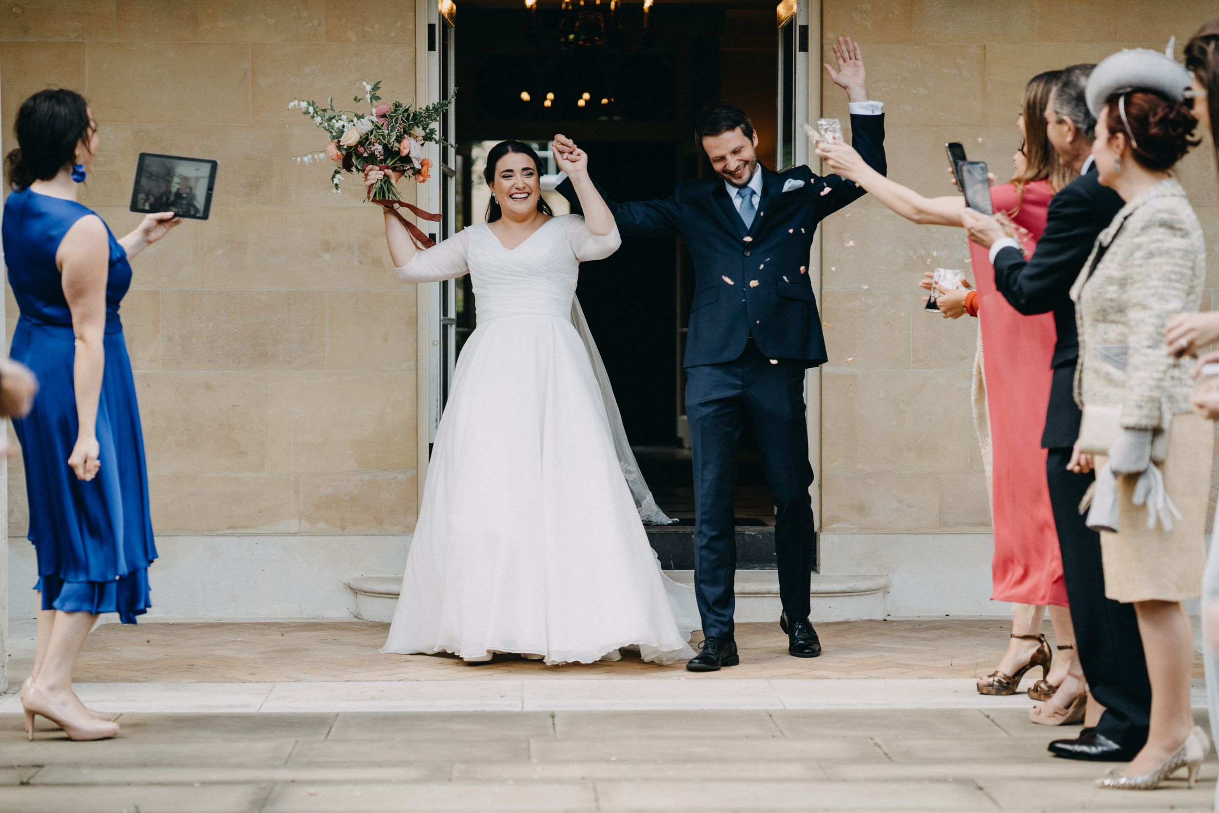 Wedding party and guests celebrating with confetti outside a traditional stone building.