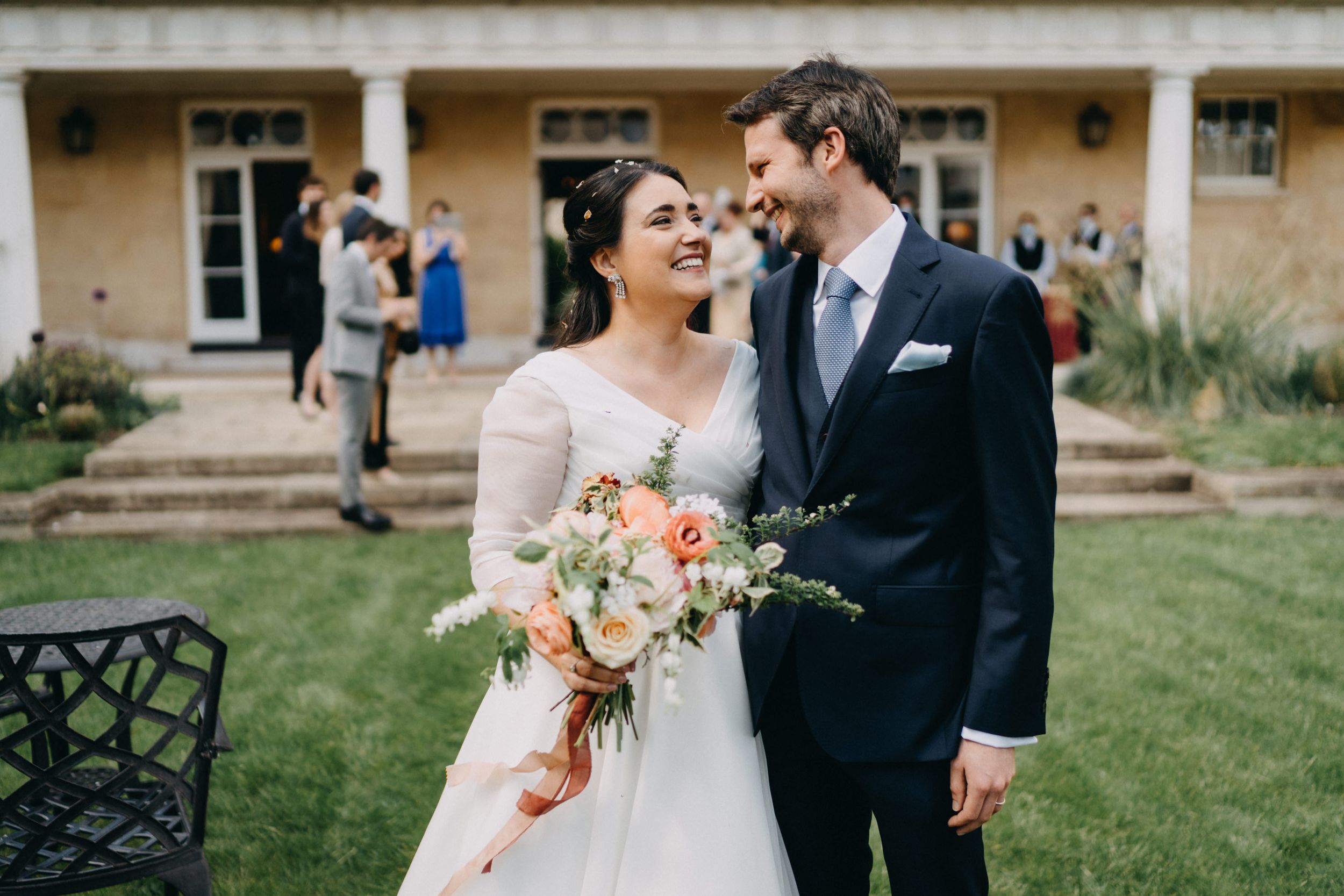 A joyful wedding couple shares a laugh while standing on a lawn with flowers at Downing College.