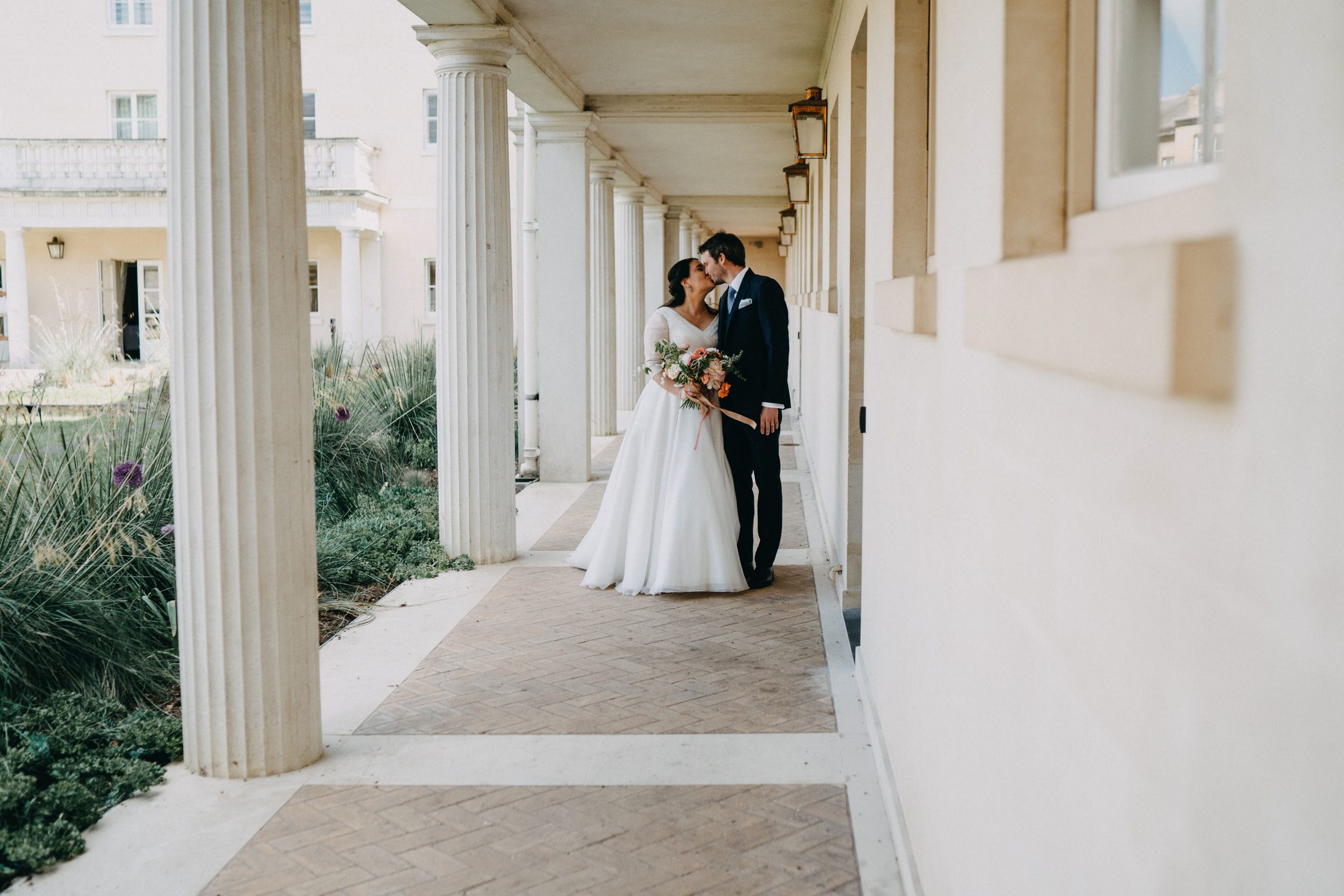 A wedding couple embraces in an elegant colonnade walkway of Downing College Cambridge.