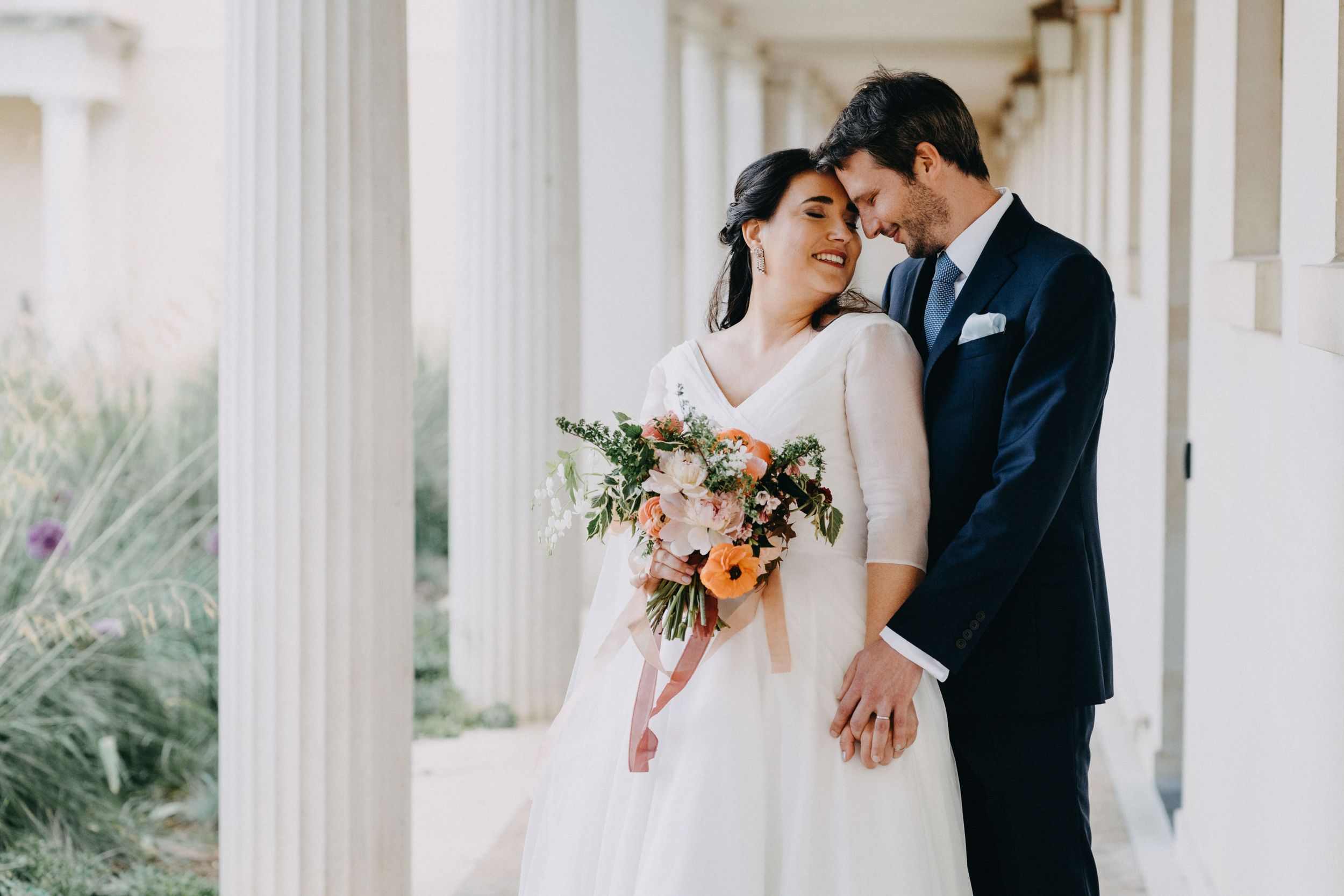 A romantic moment between newlyweds under white columns with a pink and peach bouquet.
