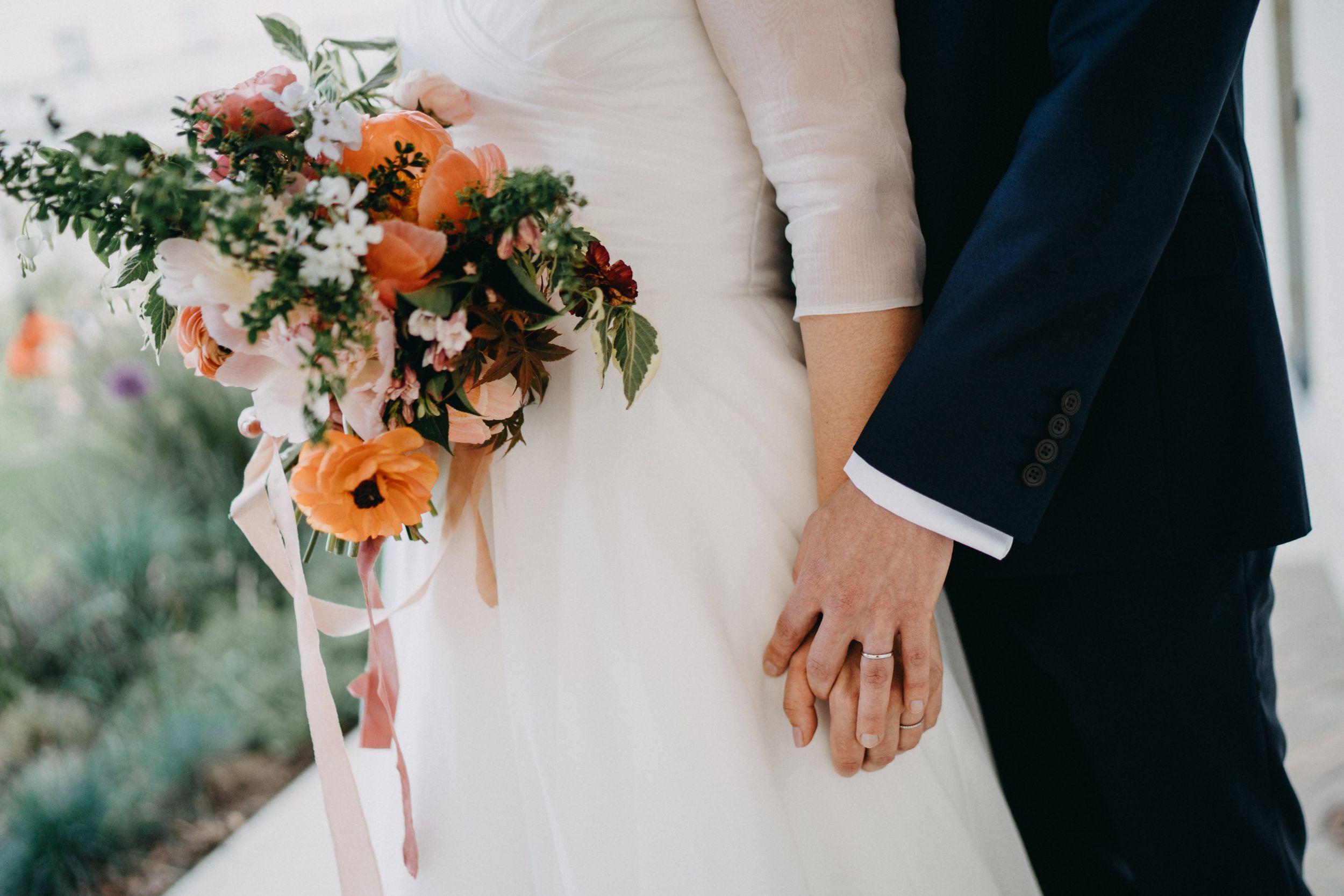 Close-up of wedding couple holding hands with vibrant orange floral bouquet.