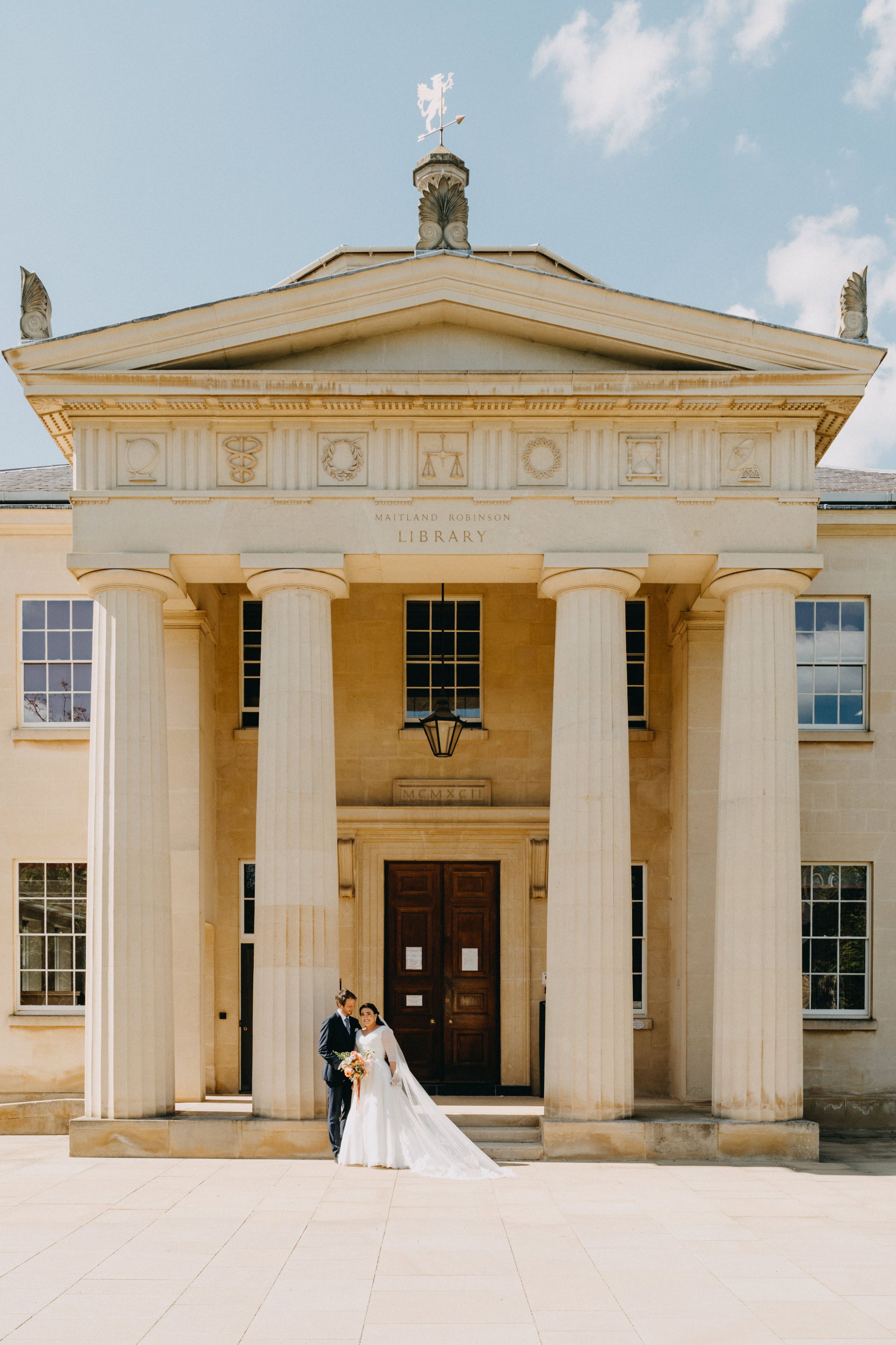 A wedding couple poses in front of a grand neoclassical building entrance with columns - Downing College Cambridge.